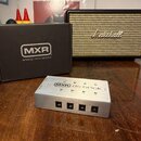 Na fotografii je stříbrný napájecí adaptér s nápisy MXR a dc brick. Zařízení má čtyři výstupní konektory a nad nimi jsou označení 9V. Vedle adaptéru…
