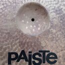 Detailní pohled na kovový činel s kovaným povrchem. Na spodní části je černý nápis PAiste a pod ním menší text MADE IN SWITZERLAND. Povrch nástroje v…