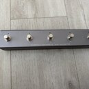 Šedý MIDI footswitch v kovovém obalu s pěti stříbrnými spínači. Na spodní hraně je bílý nápis DARKGLASS ELECTRONICS - MIDI FOOTSWITCH. Povrch zařízen…