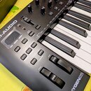 Černý MIDI kontrolér s klávesnicí. Na šasi jsou viditelná loga M-AUDIO a OXYGEN25. Nástroj obsahuje několik otočných potenciometrů, tlačítka s nápisy…