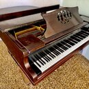 Hnědé dřevěné piano s otevřeným horním víkem, skrze které jsou vidět kovové struny a vnitřní mechanismus. Na hudebním pultu je vyřezaný kruhový ornam…