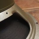 Detailní pohled na část reproduktoru s kovovým rámem stříbrné barvy a tmavou membránou. Na horní části rámu je vidět malý otvor a na povrchu membrány…