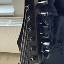 Detail hlavy černé elektrické kytary s šesti kolíky ladění. Na hraně hlavy je viditelný nápis "Ibanez". Povrch nástroje má matný černý finish a je v…