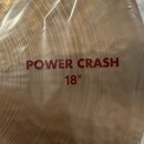 Detailní pohled na mosazný činel, který je zabalen v průhledné plastové fólii. Na povrchu je červeným písmem nápis POWER CRASH 18". Povrch činelu vyk…