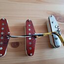 Tři elektrické snímače kytary s bílým plastovým rámečkem, položené na dřevěném povrchu. Dva levé snímače mají červeně lakované cívky s nápisem "Brami…