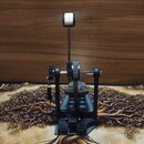 Černý bubenový pedál s kovovou konstrukcí a bílým plátnem na šlapce. Na horní části mechanismu je viditelný nápis "TAMA". Povrch nástroje vykazuje mí…