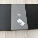 Černá obdélníková krabice s šedým papírovým obalem uprostřed. Na obalu je bílé kruhové logo s ornamentem a bílá nálepka s čárovým kódem a texty „S905…