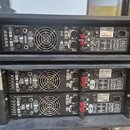 Fotografie zachycuje zadní panely tří černých rackových zesilovačů značky QSC. Na každém zařízení jsou vidět vstupy CH 1 INPUT a CH 2 INPUT, ventilát…