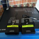 Dvě šedé bezdrátové vysílače značky SENNHEISER s nápisem XS WIRELESS. Na levém zařízení je žlutý papírek s ručně psaným textem PAVLÍK KYTARA II. a na…