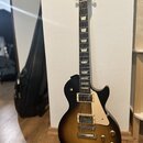Elektrická kytara s tmelelovým povrchem v přechodové barvě od černé k žluté. Na hlavě hmatníku je viditelné logo Gibson a nápis USA. Kytara má dva hu…
