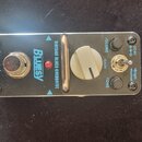 Kytarový efekt pedál v kovovém stříbrném provedení. Na povrchu jsou nápisy „TomMoye“, „BLUESY“ a „VINTAGE BLUES OVERDRIVE“. Pedál má jeden velký bílý…