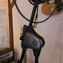 Fotografie zachycuje detail černého mikrofonního stativu s plastovou svorkou pro uchycení mikrofonu. V horní části je navinutý černý kabel s viditeln…