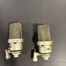 Dva kondenzátorové mikrofony stříbrné barvy s kovovými mřížkami. Na těle pravého mikrofonu je nápis -10dB 0 20dB a logo NEUMANN. Levý mikrofon má na…
