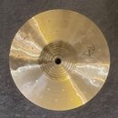 Zlatavý kovový činel s koncentrickými rýhami v centrální části. Na pravé straně je rukou napsaný nápis "Paiste". Povrch vykazuje drobné oděrky a stop…