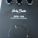 Černý kytarový výkonový zesilovač s nápisy Harley Benton, GPA-100 a Guitar Power Amplifier. Horní část obsahuje tři menší otočné ovladače s popisky B…