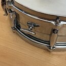 Detailní pohled na kovový snare drum stříbrné barvy s bílým membránou. Na boku korpusu jsou vidět dvě stahovací klika, z nichž na té spodní je nečite…