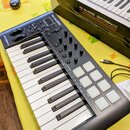 Černý MIDI kontrolér s klávesnicí, bílými a černými klávesami a osmi bílými čtvercovými pady. Na horní části je bílý nápis M-AUDIO a na levém boku ná…