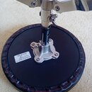 Černý kulatý sedák na chromovaném stojanu s červeným prošíváním po obvodu. Na sedáku je bílý štítek s nápisy: „K.H Musical Instruments Co., Ltd“, „Ty…