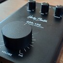 Černý výkonový zesilovač s matným povrchem. Na horní straně je velký oválný knoflík s nápisem VOLUME a označením -∞ a 0dB, dále tři menší knoflíky s…