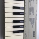 Detailní pohled na část klávesnice v šedém plastovém rámu s bílými a černými klávesami. Na pravé straně je černý nápis YAMAHA a text MIDI KEYBOARD CB…