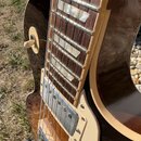 Detailní pohled na hриф a tělo elektrické kytary v tmavě hnědé barvě s krémovými plastovými díly. Hmatník z tmavého dřeva má vložky v podobě trapezoi…