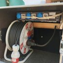 Otevřený hliníkový transportní case s černým vnitřním povrchem. Uvnitř je umístěn bílý kabelový naviják s namotaným černým kabelem a červeným konekto…