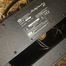 Záběr na zadní část kytarového komba s černým texturovaným povrchem. Na bílém štítku je uveden nápis Vox Amplification Ltd., Manufactured in Vietnam,…