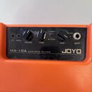 Detail ovládacího panelu oranžového kytarového komba. Na černém panelu jsou nápisy JOYO, MA-10A ACOUSTIC GUITAR a popisky ovládacích prvků: AUX IN, V…