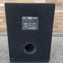 Zadní strana černého basového reproduktorového boxu s povrchem z textilie. Na horním ovládacím panelu jsou nápisy BASS CABINET, BC 210 +, 8 OHMS a 25…