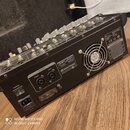 Černý míchací pult s řadou posuvníků a ovládacích prvků na horní straně. Na zadním panelu jsou vidět dva výstupy označené jako SPEAKER RIGHT a LEFT,…