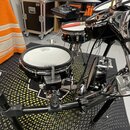 Fotografie zachycuje část elektronické bicí sady na černém stojanu. V popředí je malý bílý buben s nápisem „Roland“ a nečitelným textem nad ním. Vedl…