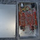 Fotografie ukazuje otevřený stříbrný kovový box s vnitřním elektronickým zapojením a oddělený kovový víko. Uvnitř je umístěna červená deska plošných…