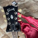 Černá hlava elektrické kytary s bílým logem Gibson a stříbrným symbolem v podobě stylizovaného krystalu. Nástroj má šest ladicích kol v krémové barvě…