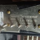 Fotografie znázorňuje zadní stranu snímače s kovovou základní deskou a černým plastovým rámem. Na desce je vyražen nápis „Seymour Duncan“ a pod ním m…