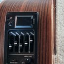 Detailní pohled na černý předzesilovač s ekvalizérem vestavěný do hnědého dřevěného korpusu akustické kytary. Panel obsahuje pět posuvníků s popisky…