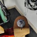 Detail konce kytarového popruhu z béžové a hnědé kůže. Na hnědém konci je kovový nýt s vyraženým nápisem „MÄNNE MAOEL“. Povrch kůže vykazuje mírné zn…