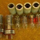 Fotografie zobrazuje skupinu elektronických vakuových elektronek a šest kovových držáků/patic. Na elektronkách jsou čitelné nápisy TUNG-SOL MADE IN R…