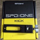 Černý kovový držák s utahovacím šroubkem z chromovaného kovu, položený na žlutočerné krabici. Na krabici jsou bílé a černé nápisy „Roland“, „SPD::ONE…