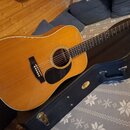 Akustická kytara s přirozenou dřevěnou barvou korpusu a černým hmatníkem. Na těle je u otvoru černý plech (pickguard) a černý kobylník. Kytara leží n…