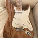 Elektrická kytara s přírodním dřevěným povrchem těla a hmatníkem z jasného dřeva. Nástroj má bílý pickguard, tři snímače typu single-coil a tři bílé…