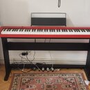 Digitální piano s červeným šasi a černými klávesami, umístěné na černém dřevěném stojanu. Nástroj má přiložený černý notový pult. Na povrchu nástroje…