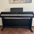 Černá digitální pianola s klaviaturou a stojanem. Na horní části je v centru bílý nápis YAMAHA. Nástroj obsahuje tři pedály a na horní desce je umíst…