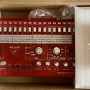 Červený elektronický bicí automat v originální krabici s polystyrenovými výplněmi. Na přístroji je bílý nápis behringer a logo s trojúhelníkem. V dol…