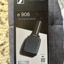 Fotografie znázorňuje originální krabici od mikrofonu. Na černé povrchové části je bílý nápis SENNHEISER s doprovodným logem, označení modelu e 906 a…