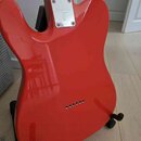 Zadní strana červené elektrické kytary s lesklým povrchem. Na krku je připevněn kovový štítek s nápisy „Larry Carlton“ a „S“. Povrch nástroje je čist…