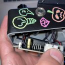 Černý efekt pedál s otevřeným spodním krytem, který odhaluje vnitřní elektroniku a zapájovanou desku. Na horním povrchu jsou ručně vymalované žluté a…