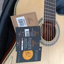 Akustická kytara s přírodním povrchem desky a hnědým mostem. Na těle nástroje jsou umístěny tři papírové štítky s logem a nápisy ORTEGA GUITARS EST.…