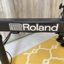 Detail černé kovové konstrukce s bílým nápisem „Roland“ a doprovodným logem. Povrch je matný a v dobrém stavu bez viditelného opotřebení. V pozadí js…