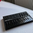 Černý kompaktní MIDI kontrolér s klávesnicí, pady a ovládacími knoflíky. Na levé straně je bílý nápis AKAI MPK mini a pod ním menší text PROFESSIONAL…