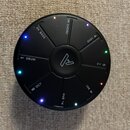 Kruhové černé elektronické zařízení s centrálním logem ve tvaru stylizovaného písmene A. Kolem středu jsou rozmístěna tlačítka s bílými nápisy: CHORD…